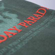 Load image into Gallery viewer, Close-up of a green Mayday Parade Comfort Colors t-shirt with red puff print text reading "PARAD," smaller partially visible white text, and a printed image on slightly folded fabric.