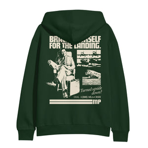 image of the back of a forest green pullover hoodie on a white background. hoodie has an image of a man sitting with a suitcase and a collage of airplanes. across the shoulders says brace yourself for the landing. on the bottom right says Turned upside down? dial 1 800 m l s c r s h