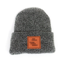 Load image into Gallery viewer, image of a black and white peppered woven winter beanie on a white background. front cuff has brown square stamped patch sewn on. five dark brown horizontal rectangles are stamped on the patch like the black lines art.
