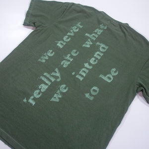 A green Mayday Parade Comfort Colors T-Shirt laid flat, featuring the text “we never really are what we intend to be” in large, faded puff print letters on the back.