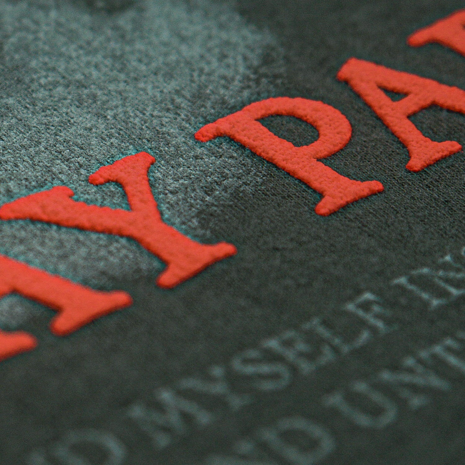 Close-up of textured black surface with large, raised red puff print letters and faint, smaller white text beneath—detail from a Mayday Parade t-shirt.