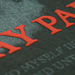 Close-up of textured black surface with large, raised red puff print letters and faint, smaller white text beneath—detail from a Mayday Parade t-shirt.