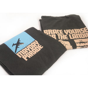 image of the front and back of a a folded pepper colored tee shirt on a white background. front is on the left and has a center chest print of a blue rectangle with an airplane. below says mayday parade. the back is on the right and has an image of a man sitting with a suitcase and a collage of airplanes. across the shoulders says brace yourself for the landing. on the bottom right says Turned upside down? dial 1 800 m l s c r s h