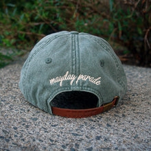 Load image into Gallery viewer, A green Mayday Parade LP101 Adams Pigment Dyed Dad Hat with a brown strap rests on concrete. &quot;Mayday Parade&quot; is embroidered in white cursive on the back, standing out among dad hats. Green foliage blurs in the background.
