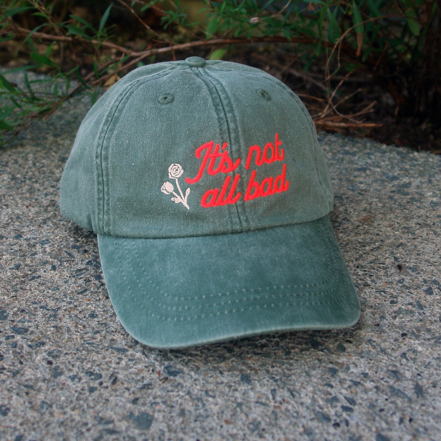 A green Mayday Parade Adams Pigment Dyed Dad Hat (Model: LP101) sits on concrete, with a white embroidered flower and red cursive "It’s not all bad" on the front. Some greenery appears in the background.