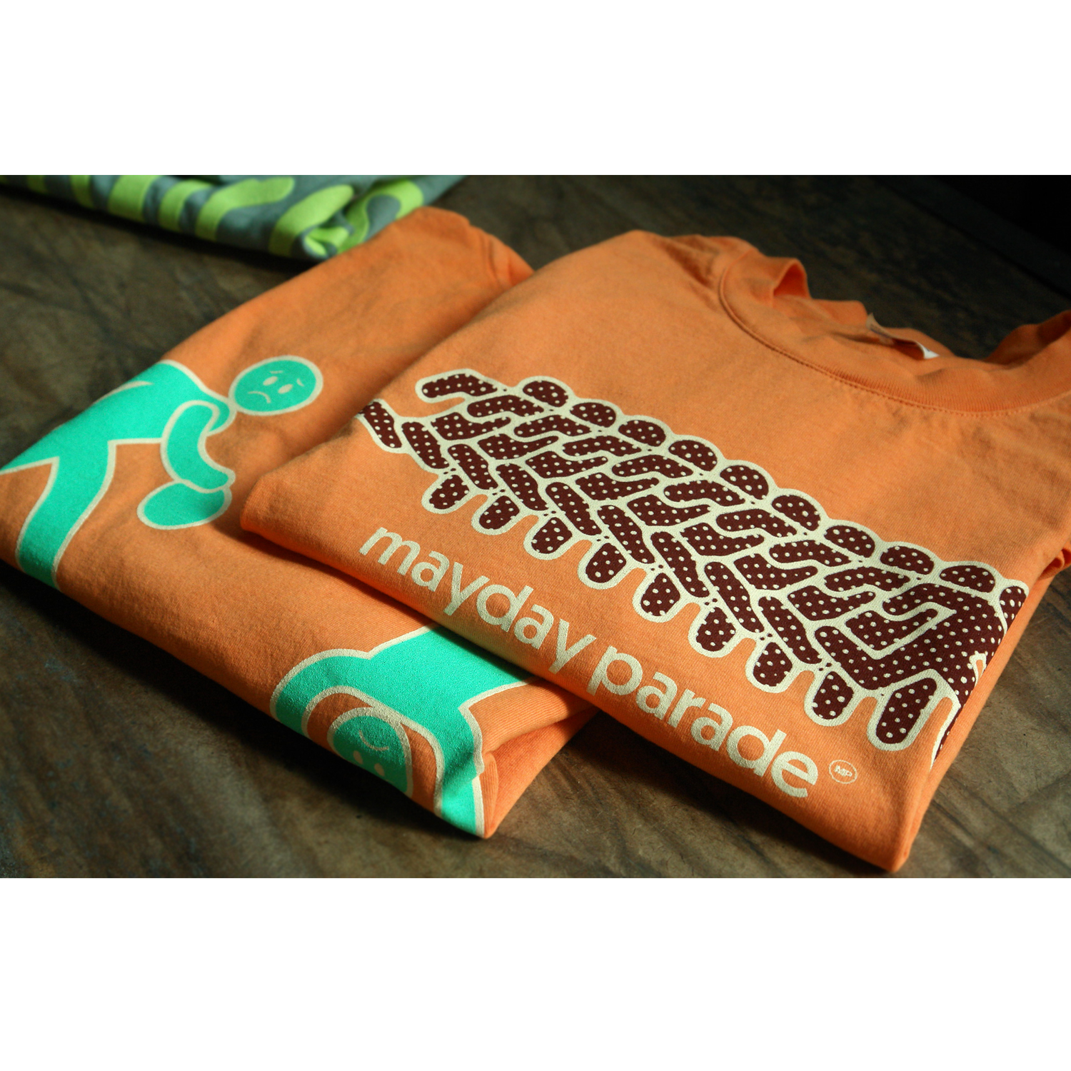 folded front and back image of a citrus orange tee shirt on a wood background. front of tee is on the right and has a running man across the front chest in maroon print and says mayday parade in white below. the back of the tee is on the left and has a full back print. in green shows a man three times, dribbling a ball, standing on his head and lifting weights. emo athleisure club is stacked in white on the bottom right of the shirt, and also says a lifestyle of sadness.