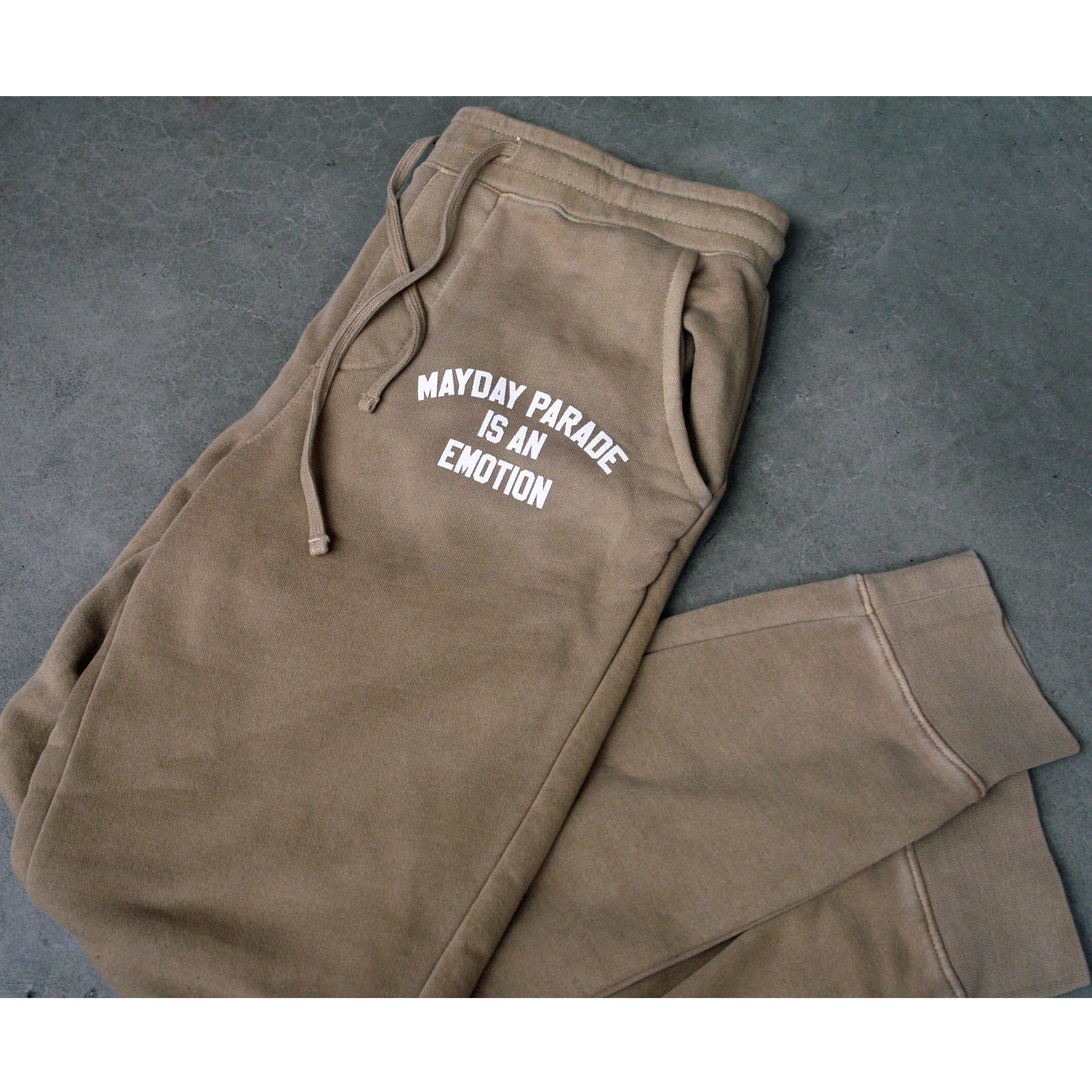 image of tan jogger sweatpants laid flat with legs folded on a concrete floor. image shows joggers drawstring laid next to the white print on top right near pocket that says mayday parade is an emotion. the legs and bottom of the joggers are shown at the bottom right of the image