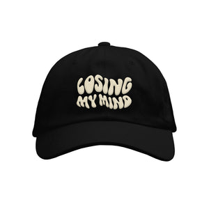 image of the front of a black dad hat on a white background. the dad hat has cream embroiderd text that says losing my mind.