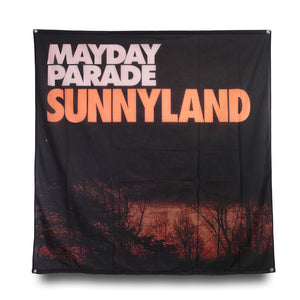 image of a square flag on a white background. flag has a full size image of a sunset with the silhouette of bare trees in front. in cream print on top left says mayday parade and in orange below across the whole flag says sunnyland