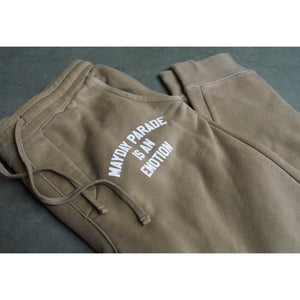 close up image of tan jogger sweatpants laid flat and folded on a concrete floor. the drawstrings of the pants lay left next to a white print near the pocket that says mayday parade is an emotion. 