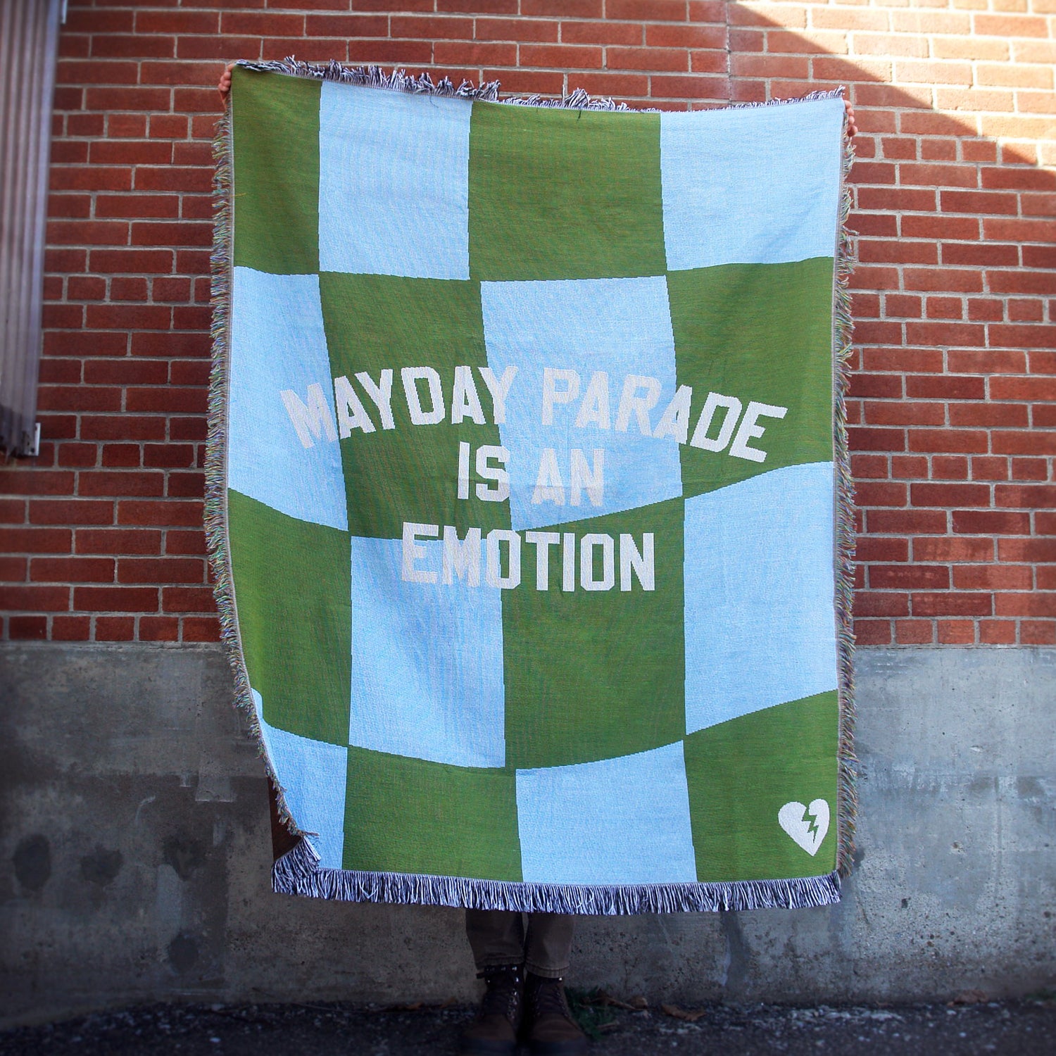 image of a custom woven blanket held up outside in front of a brick wall. blanket is green and blue checkerboard and says mayday parade is an emotion in the center.