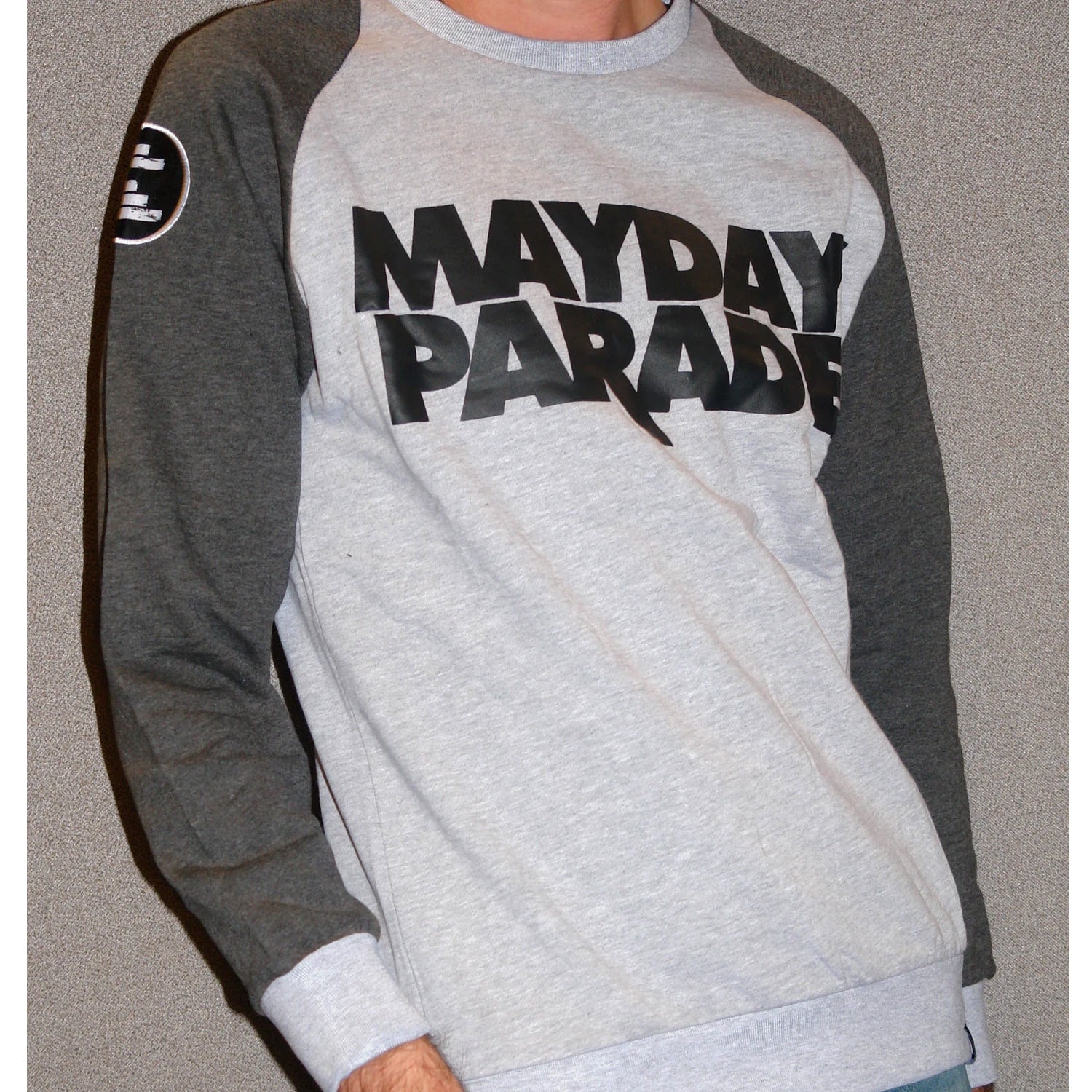 image from the neck down of a man in front of a grey background wearing a crewneck sweatshirt. the sleeves are a dark heather grey, and the chest/body is light heather grey. front chest of crewneck says stacked in black mayday parade. circle black patch with white rectangles is on the left sleeve