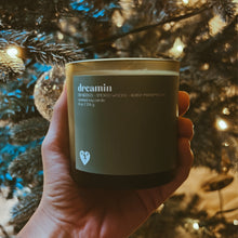 Load image into Gallery viewer, close up image of a left hand holding a candle in front of a lit Christmas tree. candle is green glass and has a teal wrapper on the front that says in white, dreamin. fir needles - smoked woods - burnt marshallow scented soy candle. 8 ounces, 226 grams. there is a small white broken heart on the bottom left with the initals M P.
