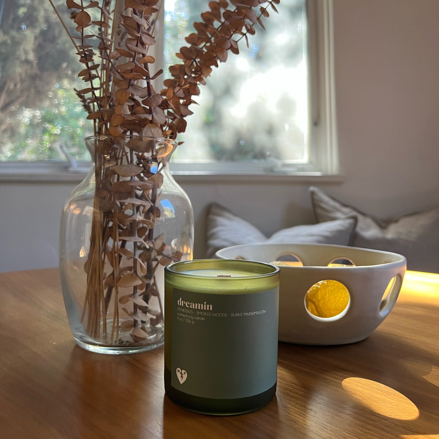 image of a table in front of a window. on the table there is a brown plant on the left in a glass vase, green candle in the center, and a bowl on the right with cut out holes and a lemon inside. 